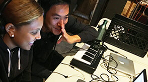 Young woman is using a computer and microphone to make beats while man is sitting in front of a keyboard.